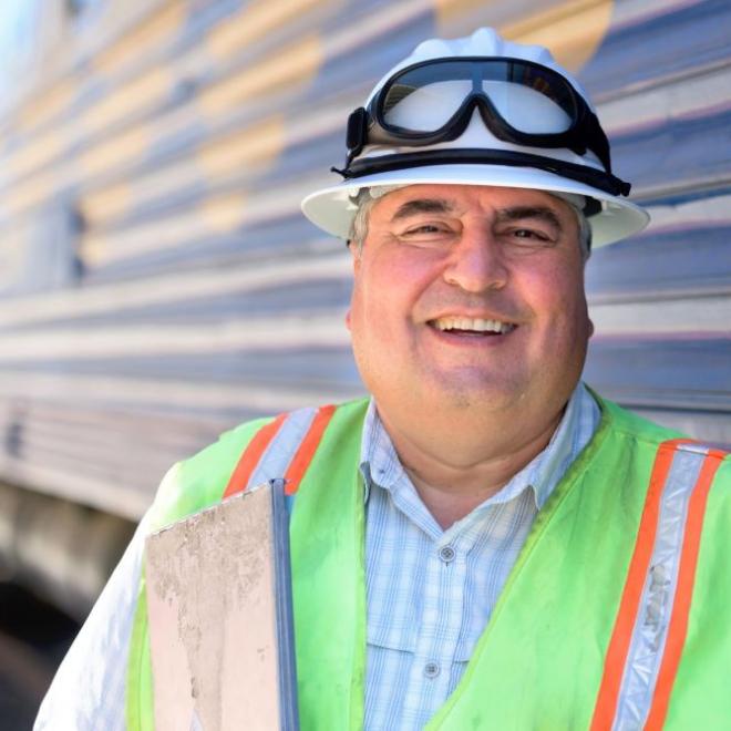 Older rail worker in high vision clothing and hard hat in front of a train.